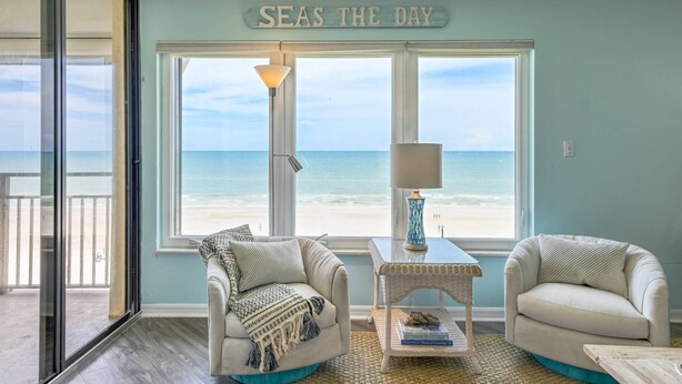 A living room with a view of the beach, a 'SEAS THE DAY' sign, and a table with a lamp and decorative items. - Sand Dollar Penthouse (St. Augustine)
