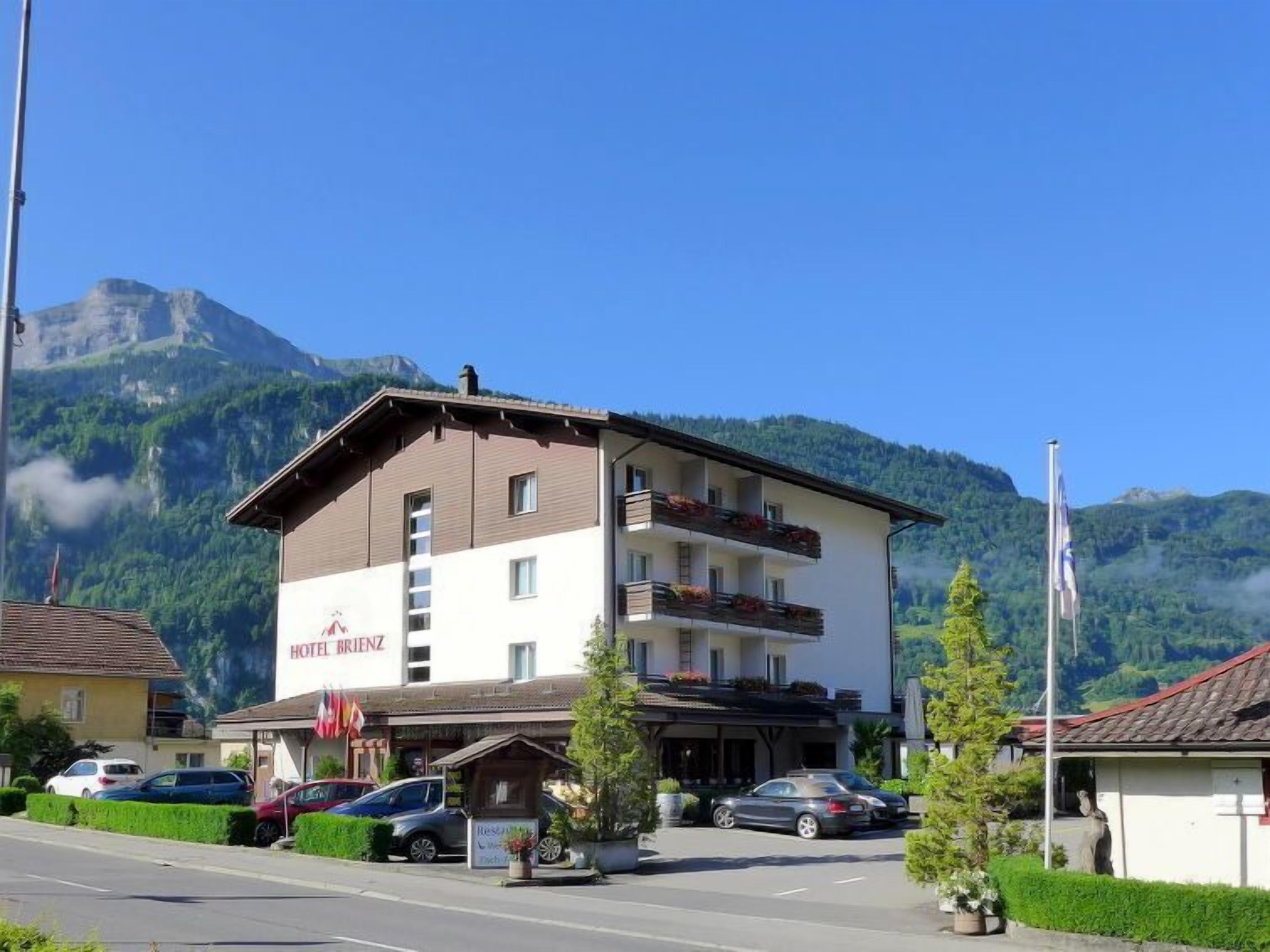 Un hôtel appelé HOTEL BRIENZ, avec une structure à deux étages, des balcons ornés de jardinières et un paysage montagneux en toile de fond.