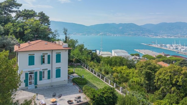 Un bâtiment à deux étages, doté d’un toit en tuiles et de volets verts, entouré d’une végétation luxuriante et offrant une vue sur une marina où sont amarrés des bateaux.