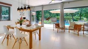 Un vaste espace dédié à la salle à manger, avec une table et des chaises en bois, un ventilateur de plafond et de grandes fenêtres offrant une vue sur une piscine et un jardin luxuriant.