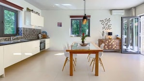 Une cuisine moderne avec des meubles blancs, un coin repas doté d’une table en verre et une vue sur la verdure à l’extérieur.