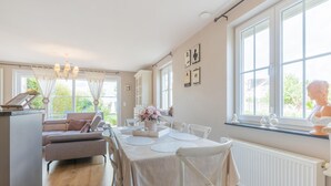 A dining area with a table set for four, white chairs, a chandelier, and a window with a view of greenery. - Inviting holiday home in Nieuwpoort with garden (Nieuwpoort)