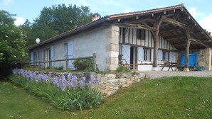 Uma casa rústica de pedra com varanda de madeira, um muro de pedra e um jardim com flores roxas.