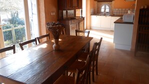 Uma mesa de jantar de madeira com cadeiras, um vaso com flores secas e vista para uma cozinha com armários brancos e painel de azulejos.