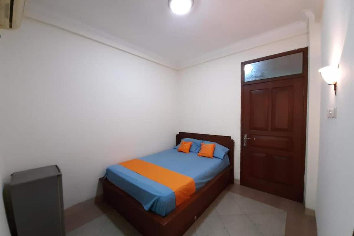 Ein kleines Einzelzimmer mit einem Holzbettgestell, einer blauen und orangefarbenen Bettdecke, einer kleinen Mülltonne und einer Holztür mit einem Spiegel darüber.
