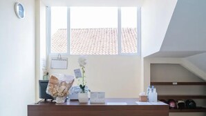 A reception desk with a potted plant and a small vase, a window with a view of a tiled roof, and a shelf with hats.