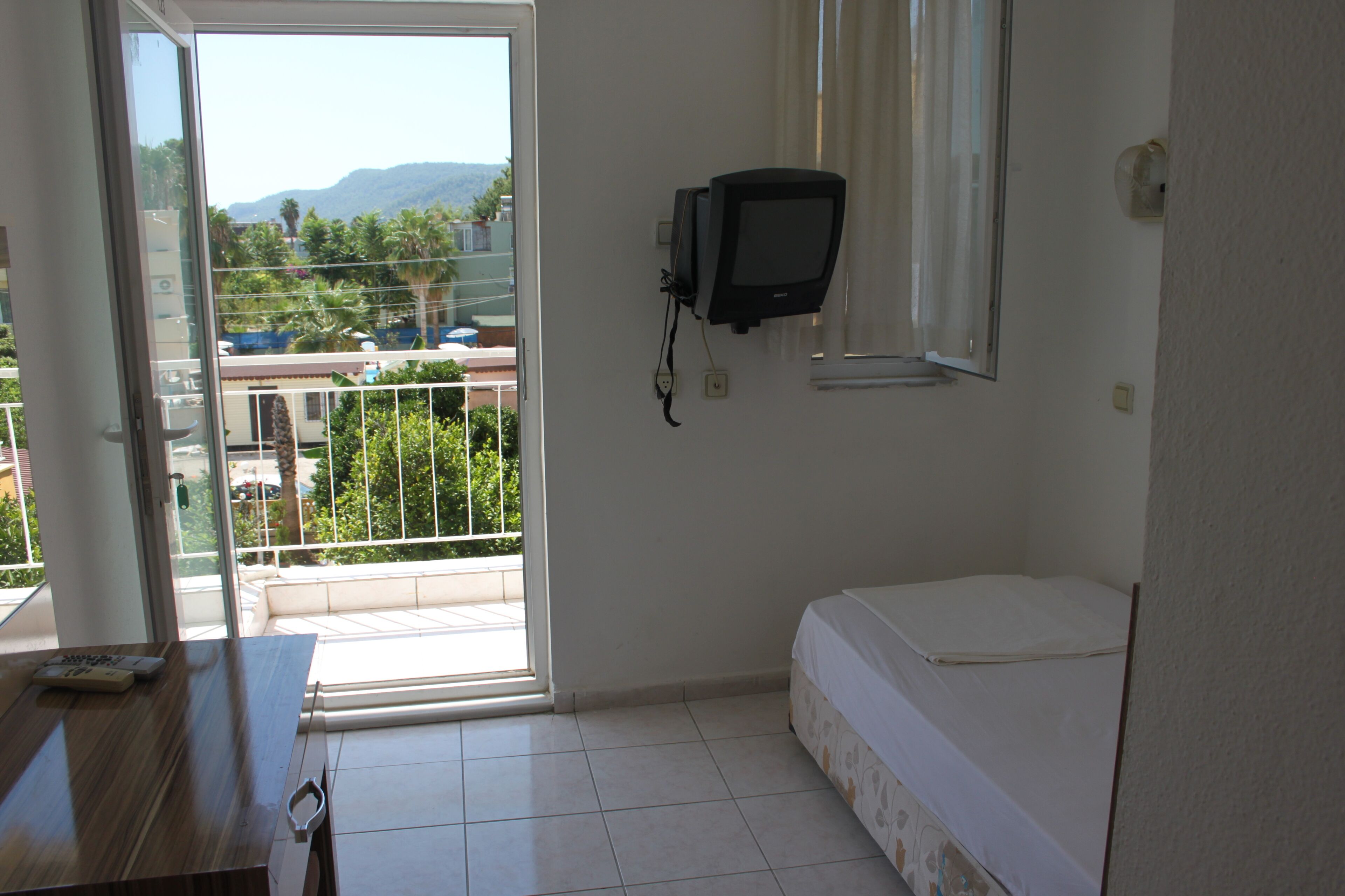 A room with a bed, a television mounted on the wall, a window with a view of greenery and buildings, and a door leading to an outdoor area.