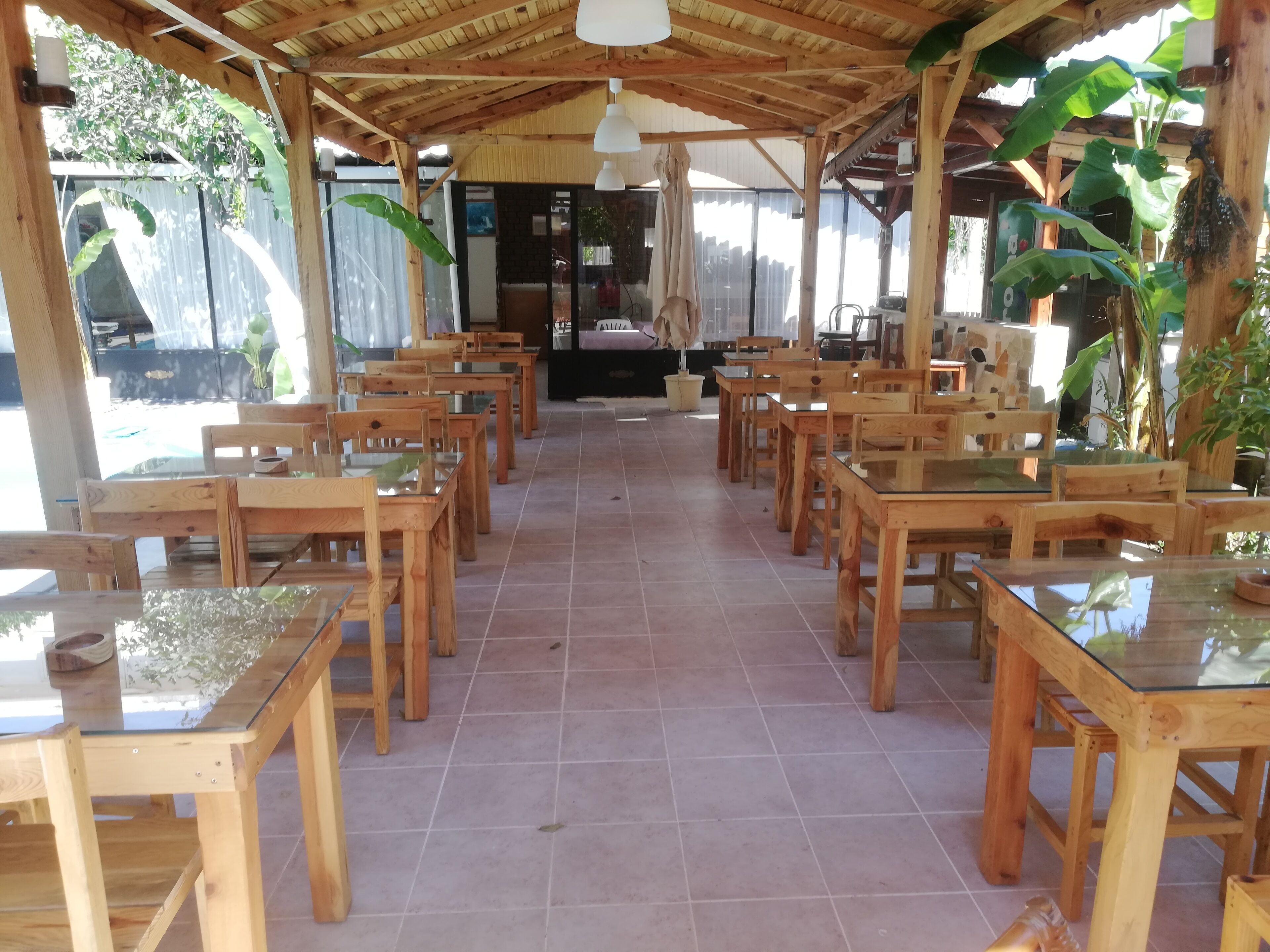 A covered outdoor dining area with wooden tables and chairs, potted plants, and a view of the surrounding area.
