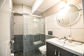 A modern bathroom with a glass-enclosed shower, a white sink with a mirror, and toiletries. - İrinikos Otel (Bozcaada)