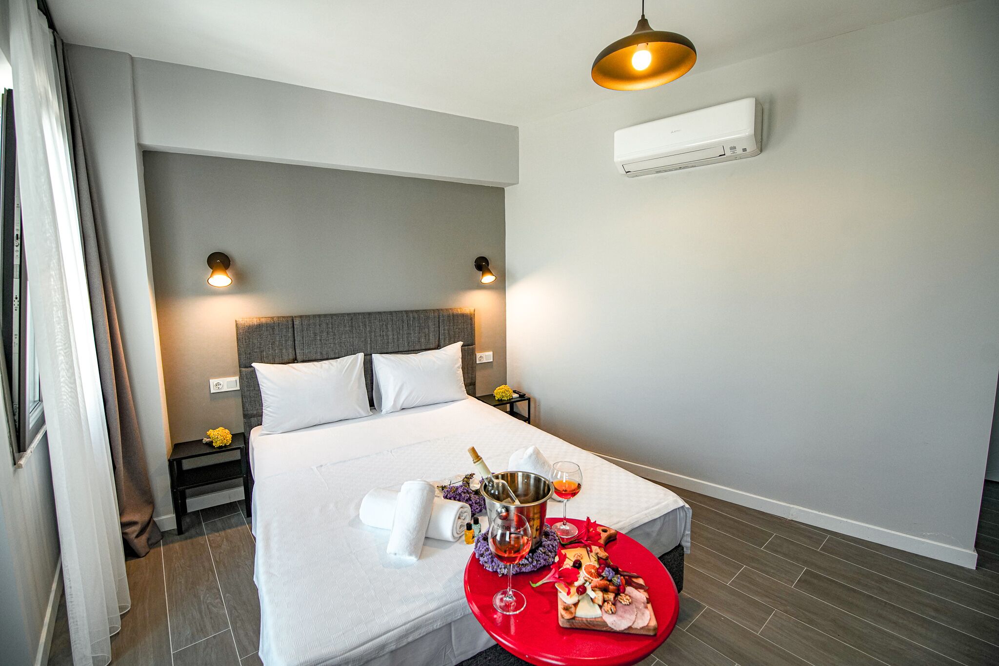 Een hotelkamer met een opgemaakt bed, een nachtkastje met een fles en glazen, en een rode dienblad met eten en een drankje.