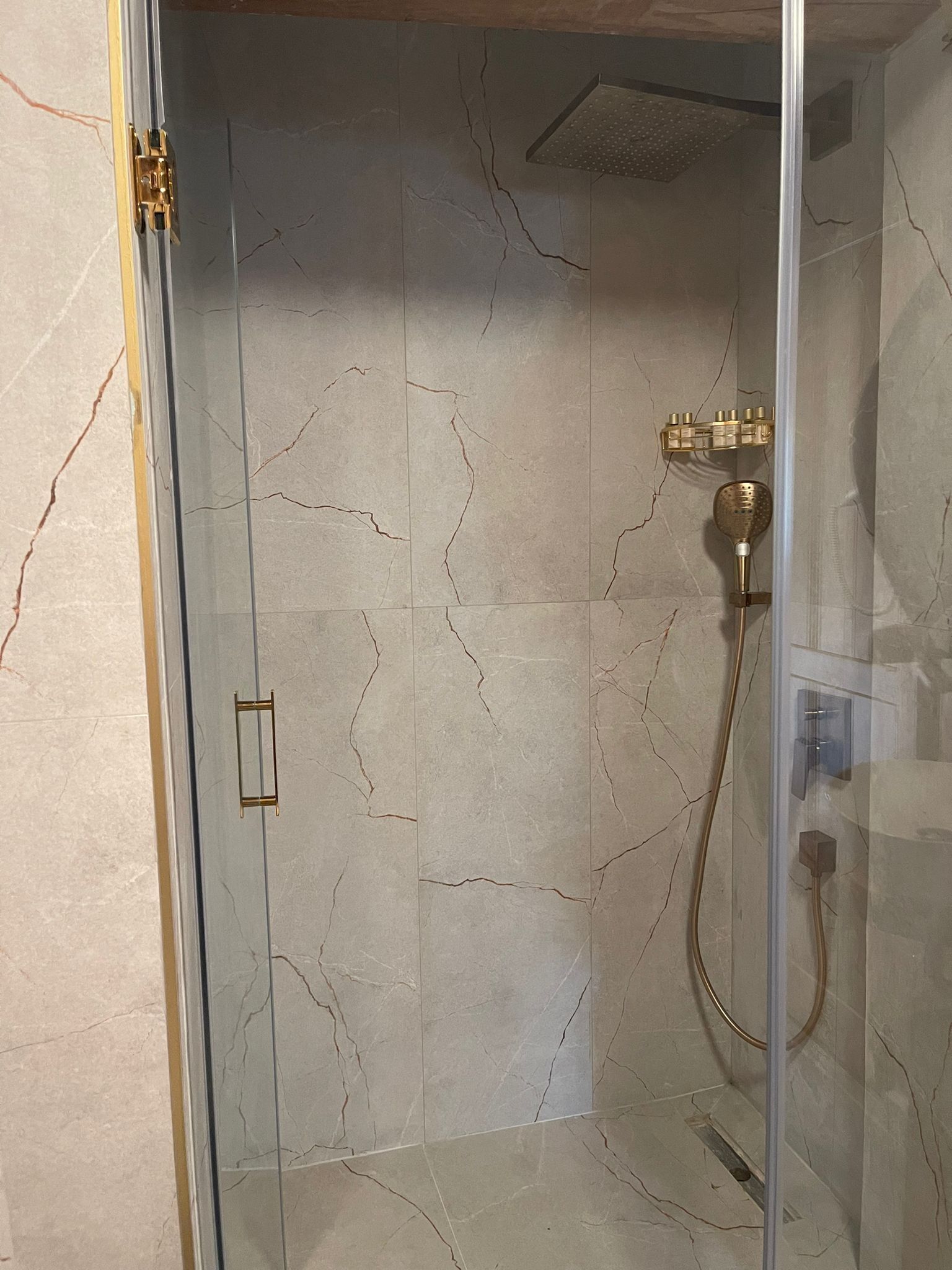 A glass-enclosed shower with a marble wall, a gold faucet, and a handheld showerhead.