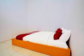 Una cama con una colcha blanca y roja, un almohadón rojo y un almohadón blanco frente a una pared lisa.