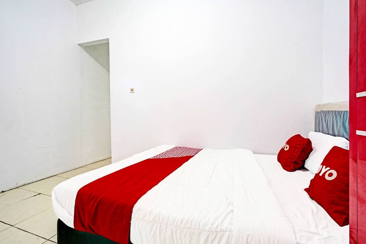 Un lit recouvert d’une couvre-lit rouge et blanche, deux oreillers rouges ornés du mot « YO », et un mur blanc avec une petite fenêtre.