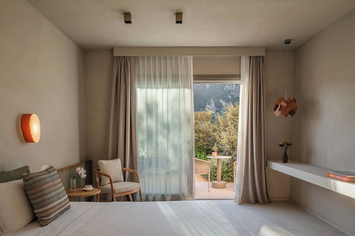 Ein Hotelzimmer mit einem Bett, einem Sessel, einem kleinen Tisch, einer Vase mit Blumen und Blick auf Grünfläche.