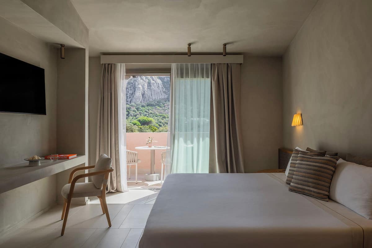 Ein Hotelzimmer mit einem großen Bett, einem Schreibtisch mit Stuhl, einem Fernseher und Blick auf eine gebirgige Landschaft durch ein Fenster mit Vorhängen.