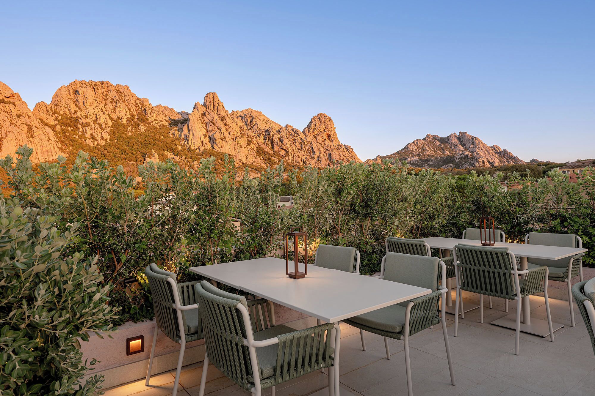 An outdoor dining area with tables and chairs, surrounded by greenery and overlooking rocky mountains.