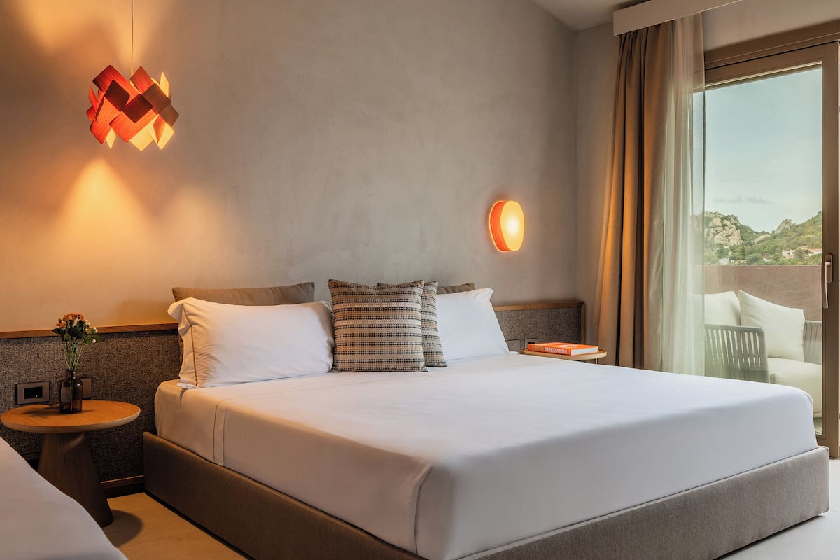 Ein Hotelzimmer mit einem großen Bett, einem Nachttisch mit Blumenvase, einer Wandlampe und Blick ins Freie durch ein großes Fenster.