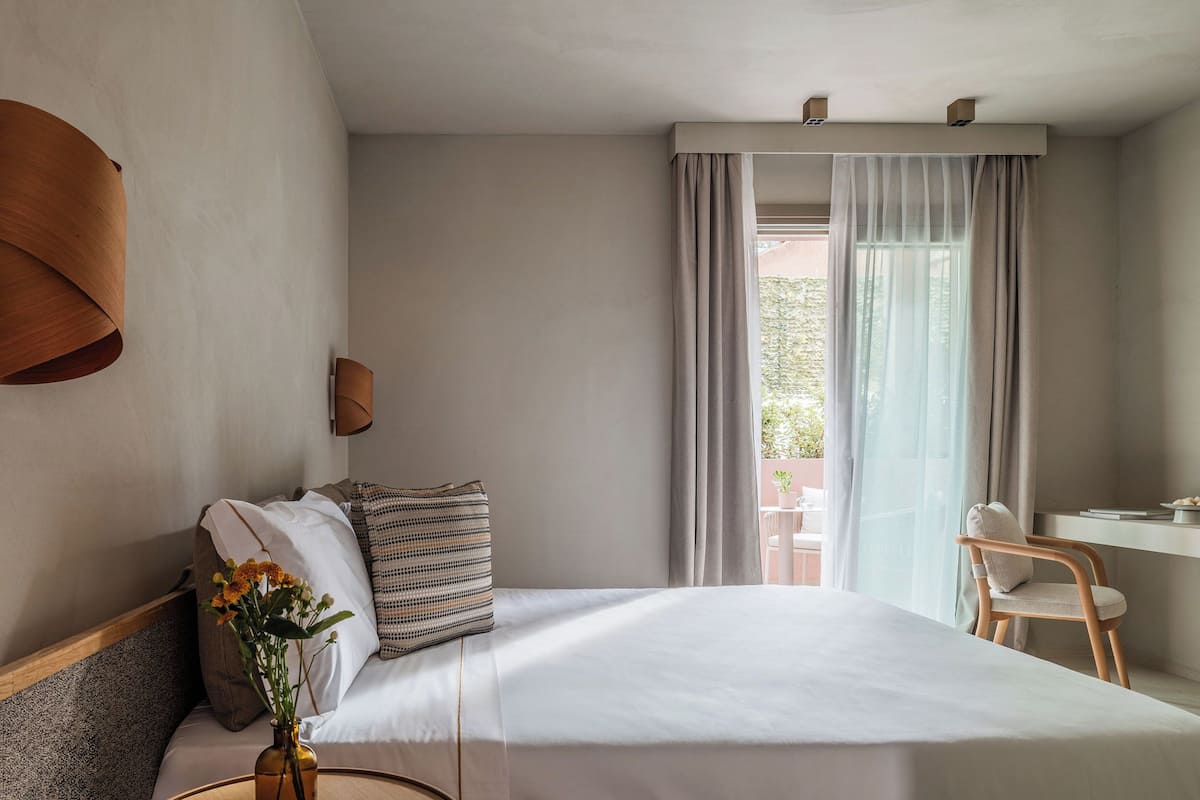 Ein Hotelzimmer mit Bett, Schreibtisch, Stuhl, einer Vase mit Blumen und einem Fenster mit Vorhängen.