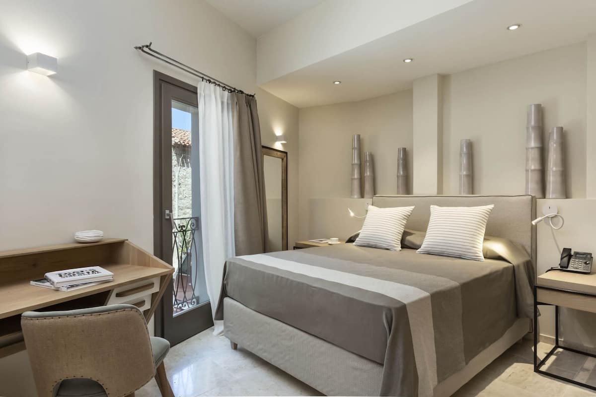 Ein modernes Hotelzimmer mit einem großen Bett, einem Schreibtisch mit Stuhl, einem Nachttisch mit Lampe und Blick ins Freie.