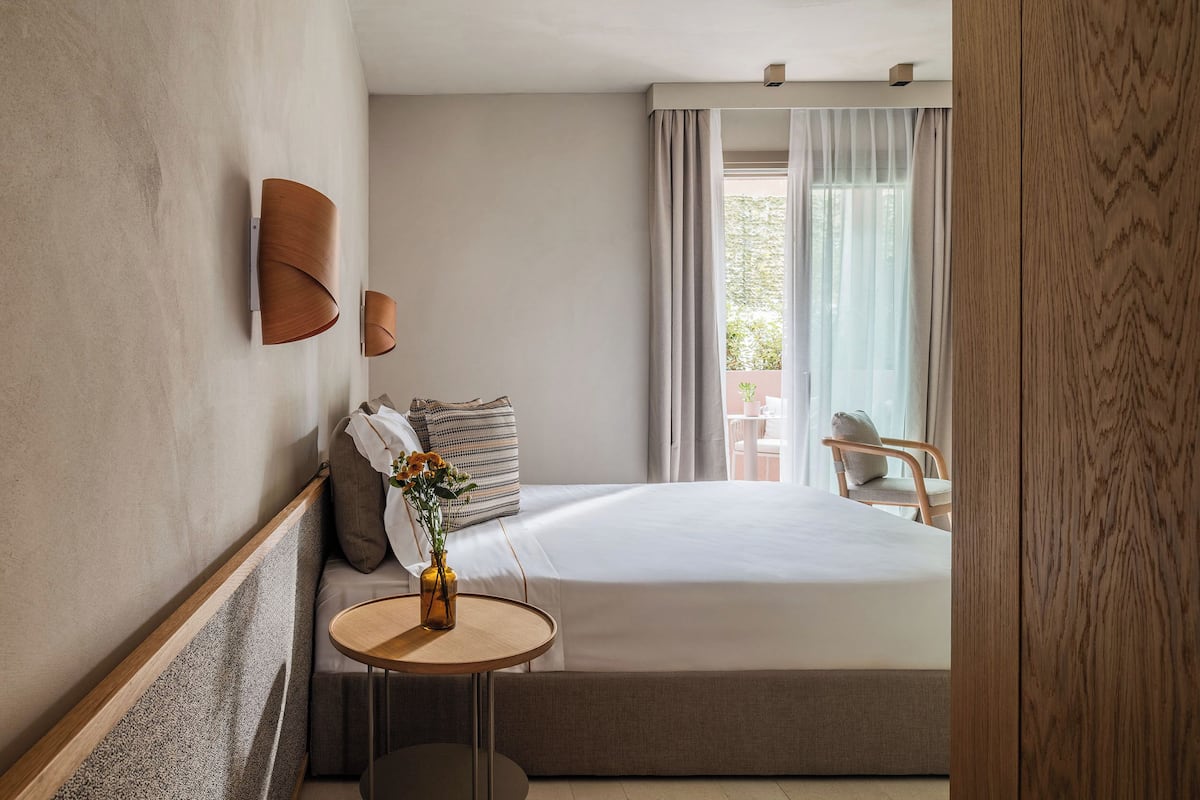 Ein Hotelzimmer mit Bett, einem kleinen runden Tisch mit Vase, einer Wandlampe und Blick ins Freie durch ein Fenster mit Vorhängen.