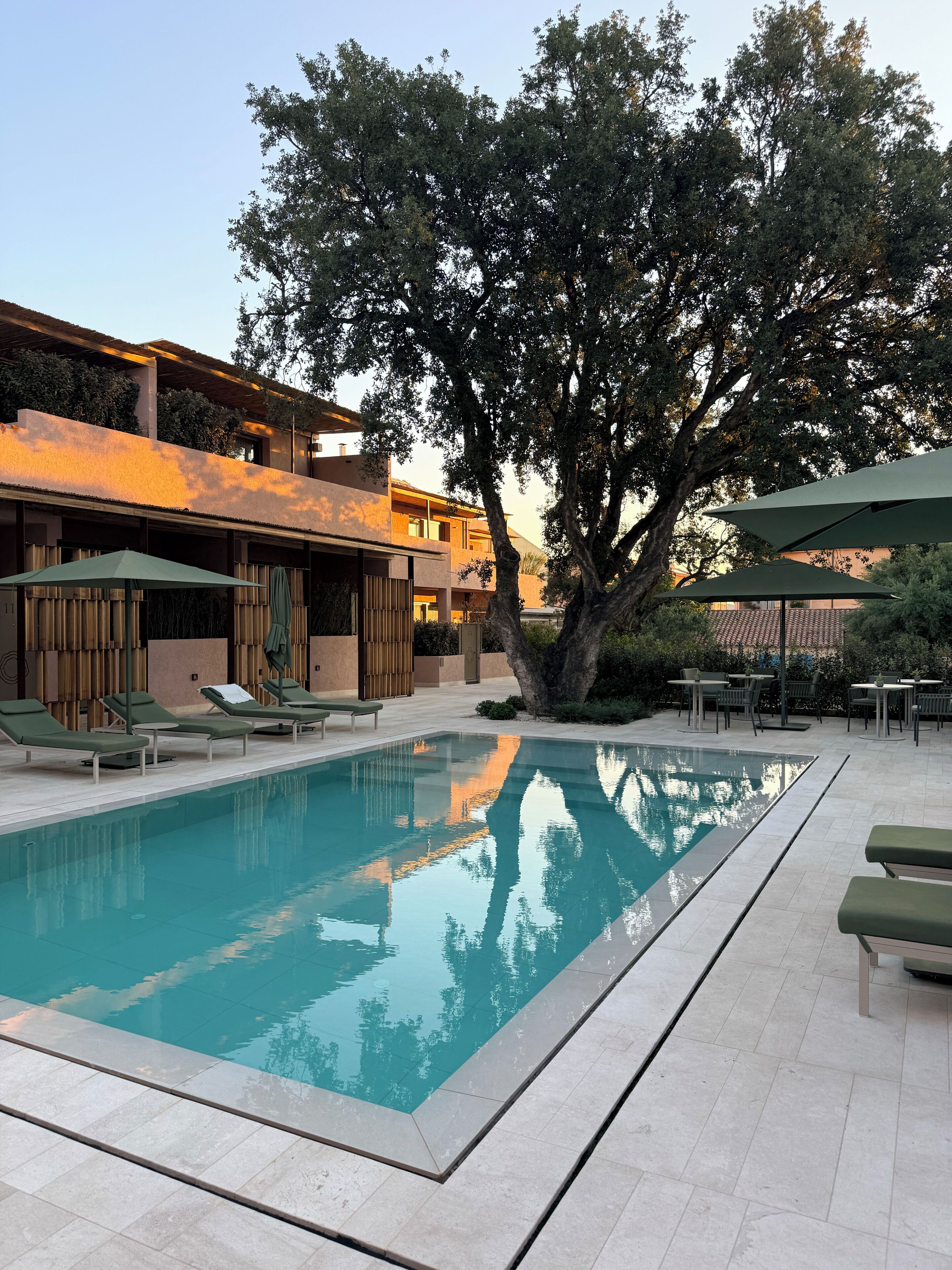 Una piscina con sillas de descanso y sombrillas, rodeada de árboles y un edificio.