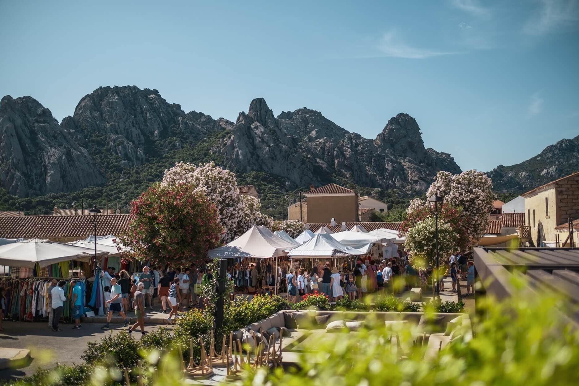 A market area with white tents and people, surrounded by buildings and mountains.