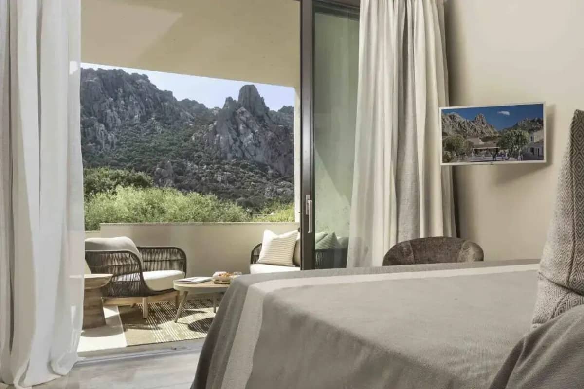 Ein Hotelzimmer mit Bett, kleinem Tisch, Stuhl und Balkon mit Blick auf felsige Berge.