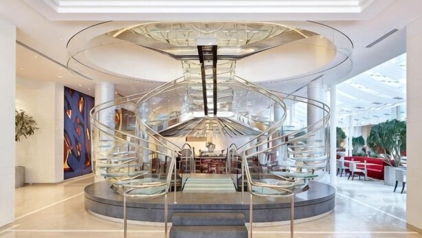 A modern hotel lobby with a central spiral staircase, a large glass ceiling, and a seating area with tables and chairs.