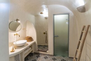 A bathroom with a white sink, a round mirror, a shower with a glass door, and patterned floor tiles. - Afora Houses (Santorini)