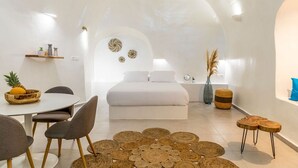 Un dormitorio moderno y minimalista con una cama grande, una zona de comedor con una mesa redonda y sillas, y un pequeño rincón de descanso con un taburete.