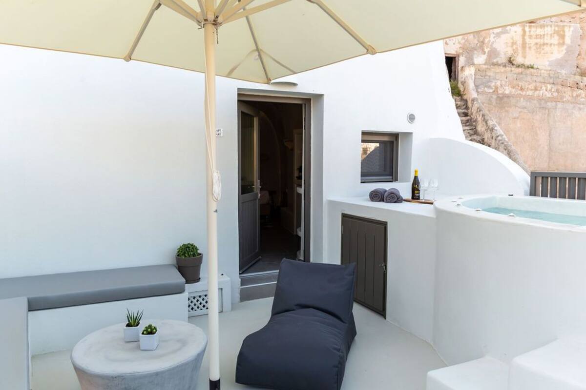 Un área de estar exterior moderna con una silla lounge, una mesita redonda con plantas, un jacuzzi y una puerta que da a un espacio interior.
