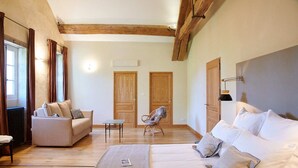 A spacious room with a large bed, a sofa, a chair, and a small table. There are wooden beams on the ceiling and a window with curtains.