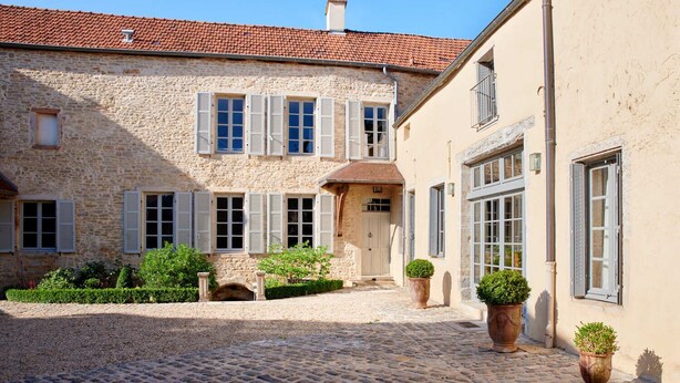 A courtyard with cobblestone pavement, stone buildings with shutters, and potted plants.