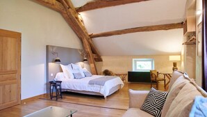A cozy bedroom with a bed, a sitting area with a couch and chairs, wooden beams on the ceiling, and a window providing natural light.