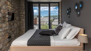 A bedroom with a stone wall, a bed with white and patterned bedding, and a view of buildings through large windows.