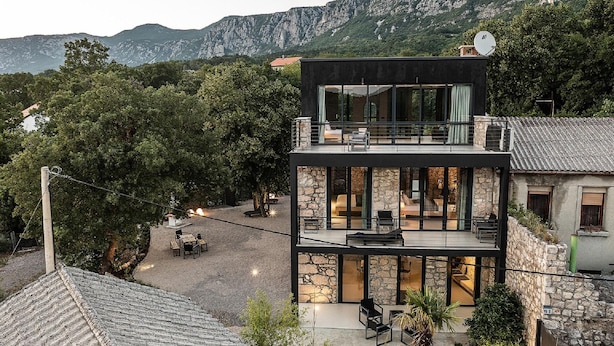 A modern two-story house with a stone facade, large glass windows, and a balcony overlooking a mountainous landscape.