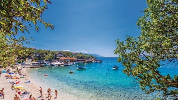 A beach with clear turquoise water, people sunbathing, and boats docked near the shore.