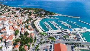 An aerial view of a coastal town with a marina, buildings with red roofs, and a clear blue sea.