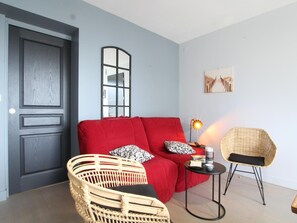 Une pièce avec une porte en bois foncé, un canapé rouge orné de coussins à motifs, un fauteuil en osier, une petite table avec une lampe et un tableau accroché au mur.