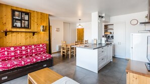 A modern apartment with a living area featuring a red patterned sofa, a wooden coffee table, and a dining area with a wooden table and chairs. The kitchen has white cabinets, a central island, and a refrigerator. - Modern 4-room apartment, 2 terraces, covered parking, ski-in/ski-out (La Plagne Montchavin Les Coches)