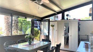 En overdækket terrasse med et glasbord og stole, en ventilator i loftet og udsigt til grøn vegetation.