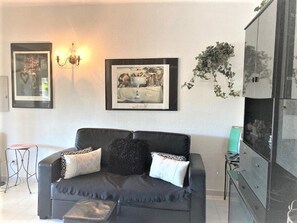 Un salon avec un canapé gris foncé, deux coussins décoratifs à motifs, une œuvre d’art en noir et blanc encadrée, un luminaire fixé au mur et une plante en pot.