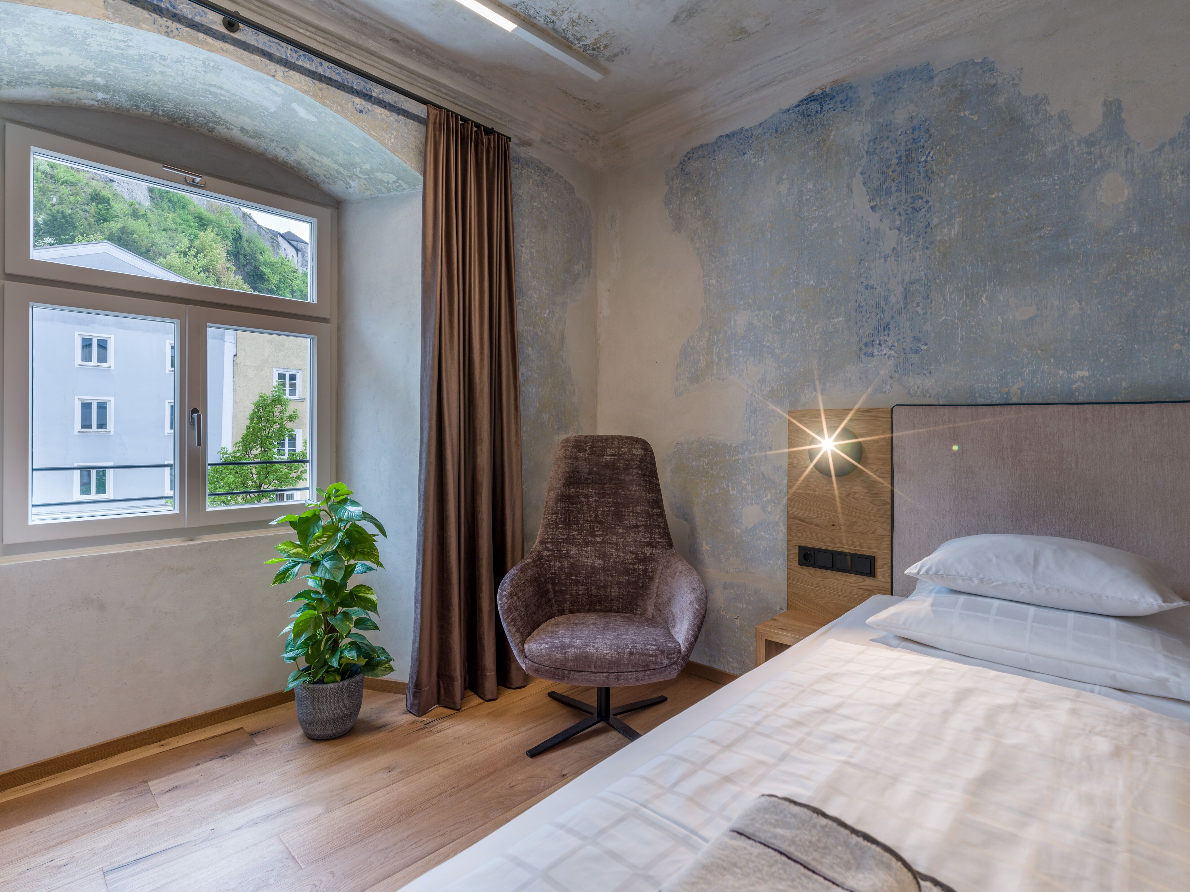 A bedroom with a bed, a chair, a potted plant, and a window with a view of buildings and greenery.