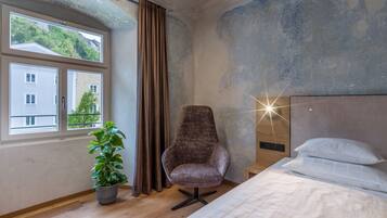 A bedroom with a bed, a chair, a potted plant, and a window with a view of buildings and greenery.