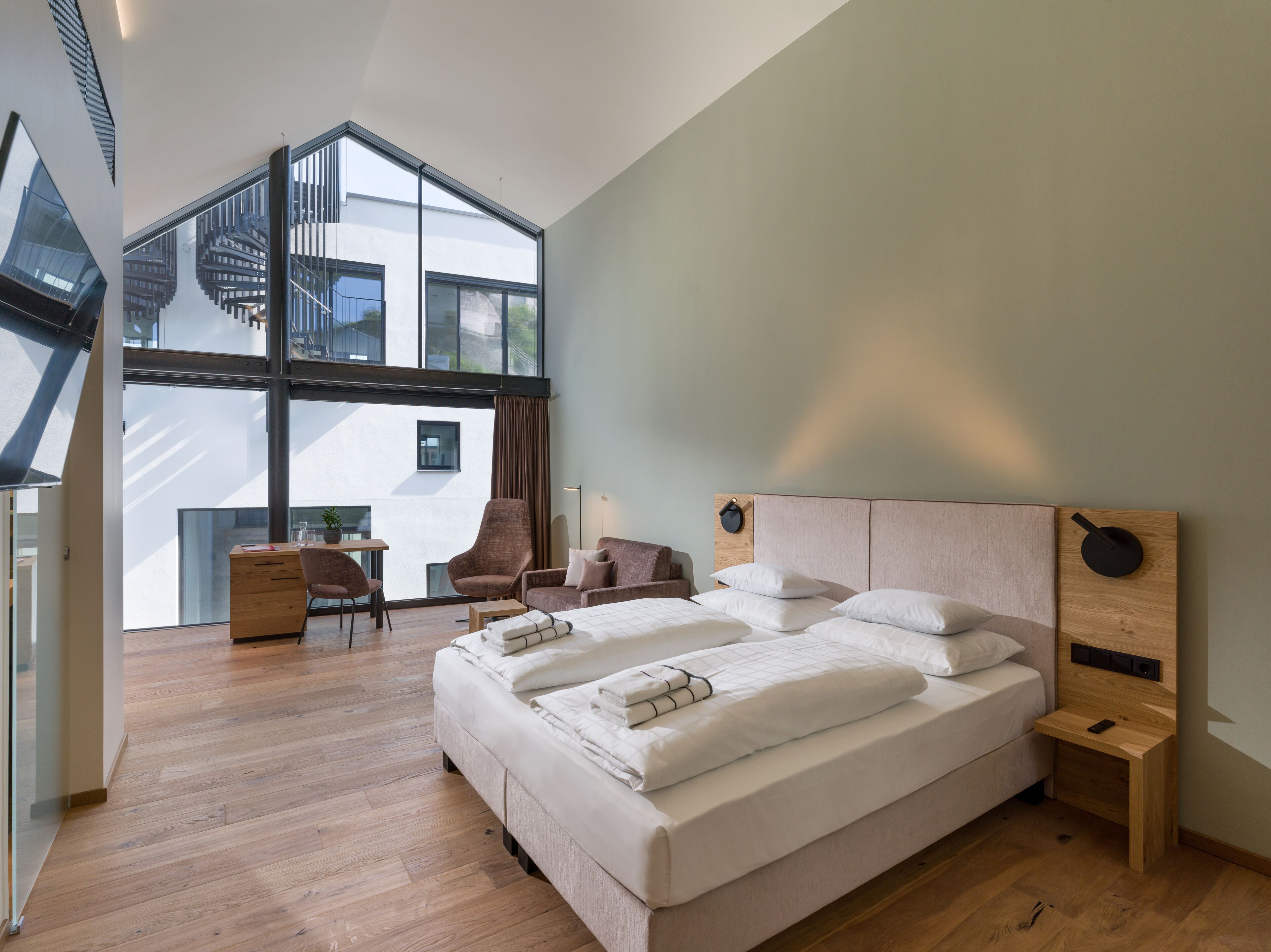 A modern hotel room with a large bed, a desk, and a chair. There is a view of the city through large windows.