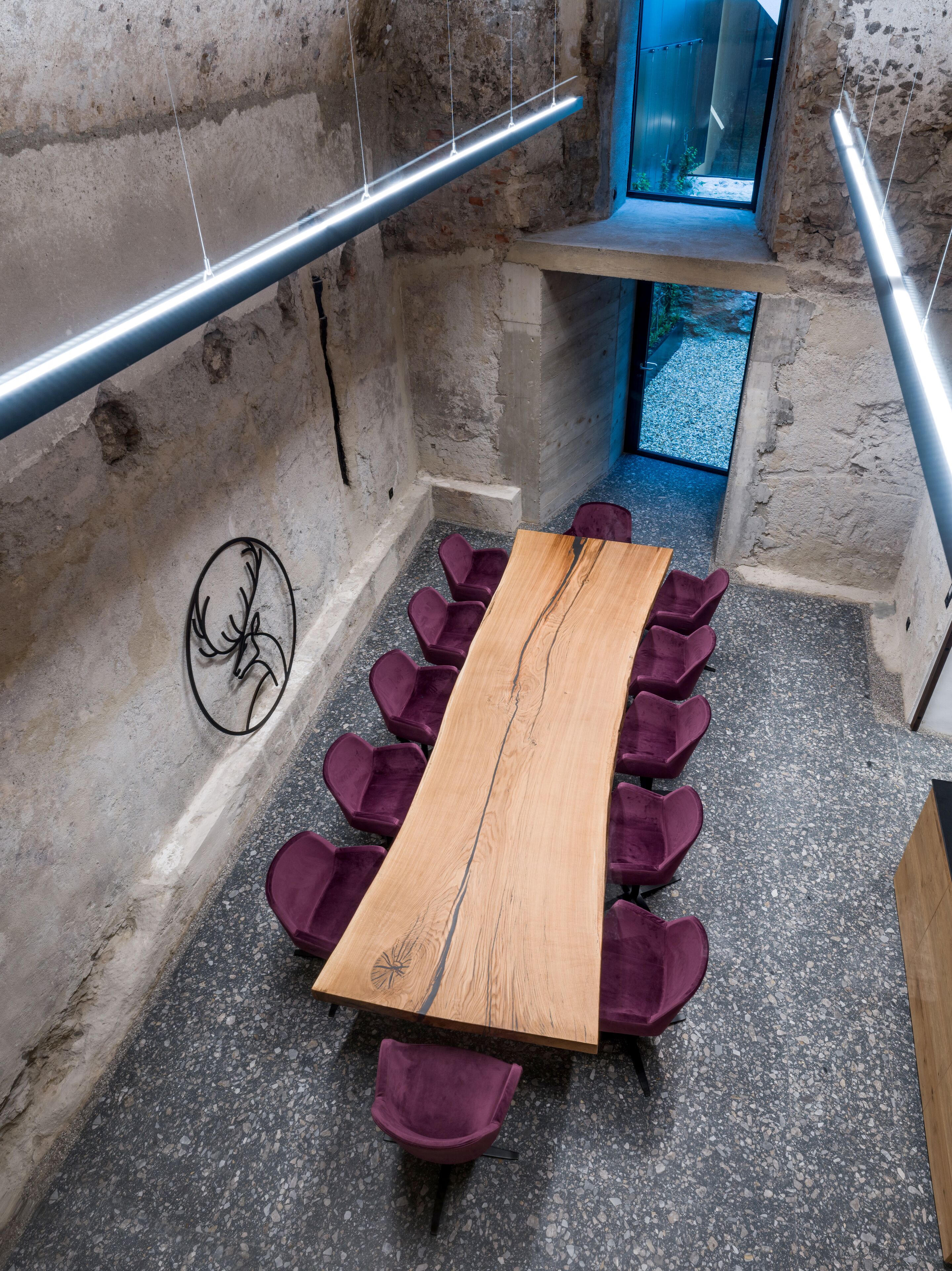 Un salón de conferencias con una larga mesa de madera y sillas moradas, paredes de piedra y una barandilla metálica.