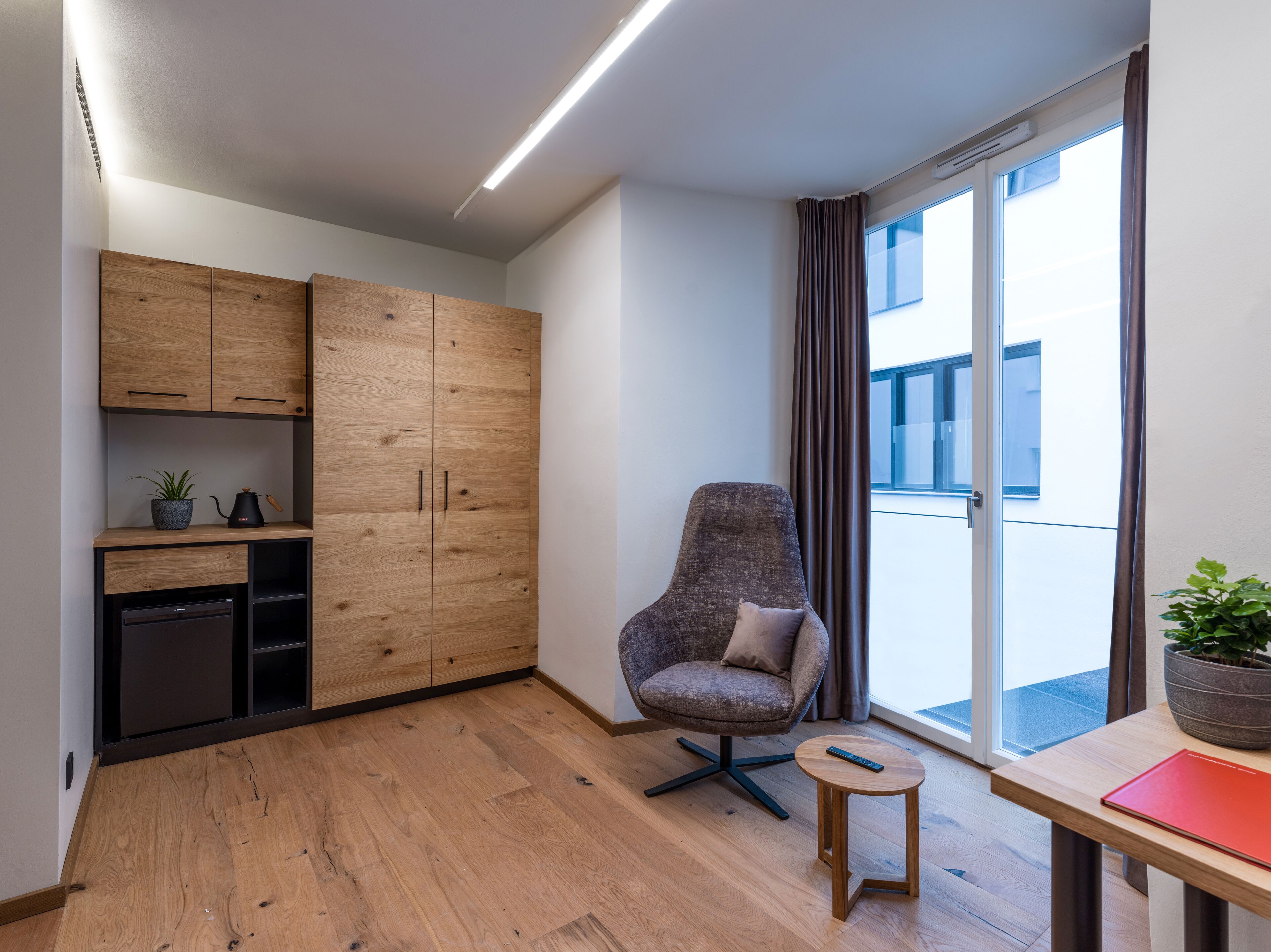 A modern room with wooden cabinets, a grey armchair, a small table, and a large window with curtains.