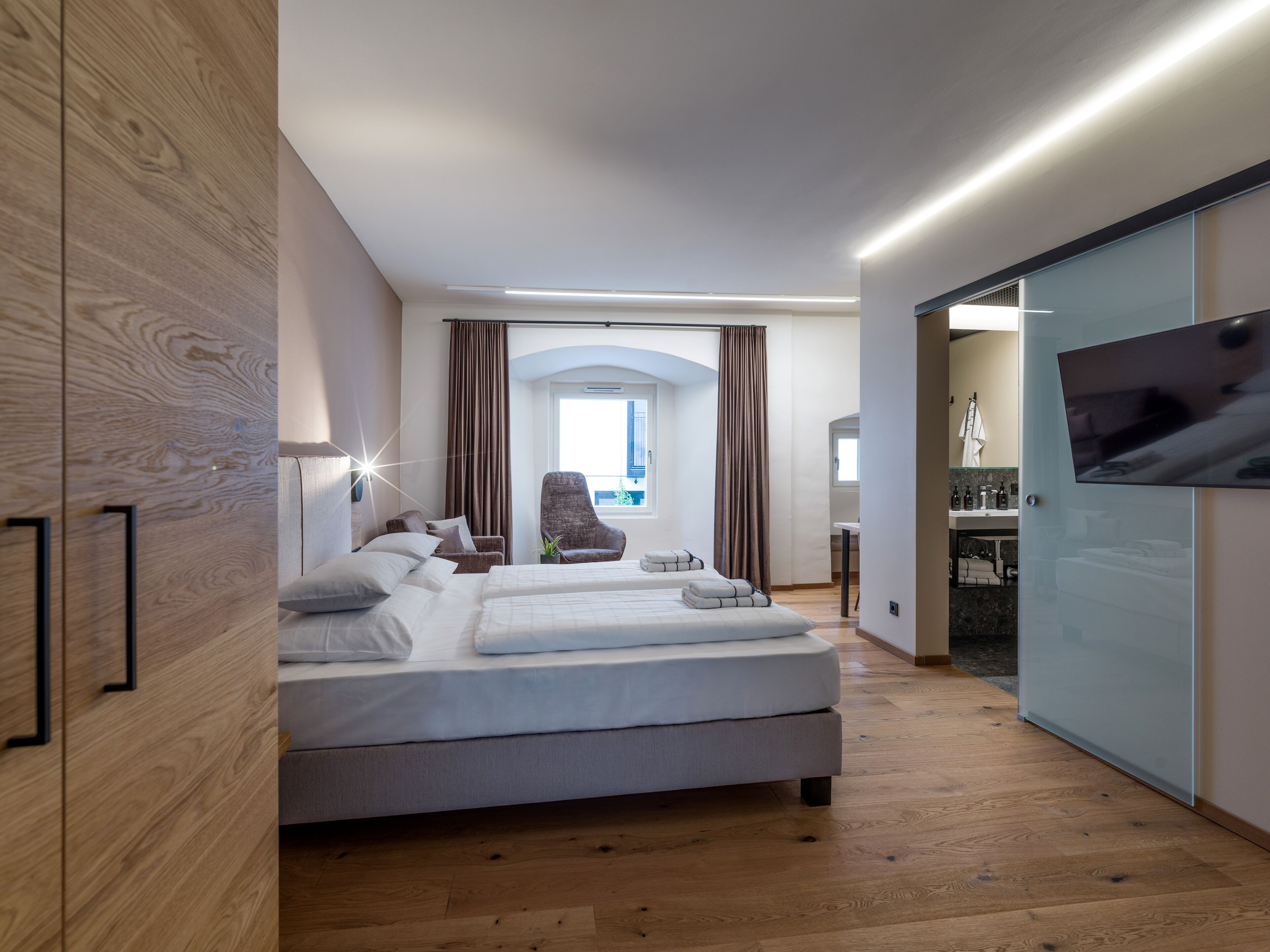 A modern hotel room with a large bed, a chair, a TV, and a bathroom visible through an open door.