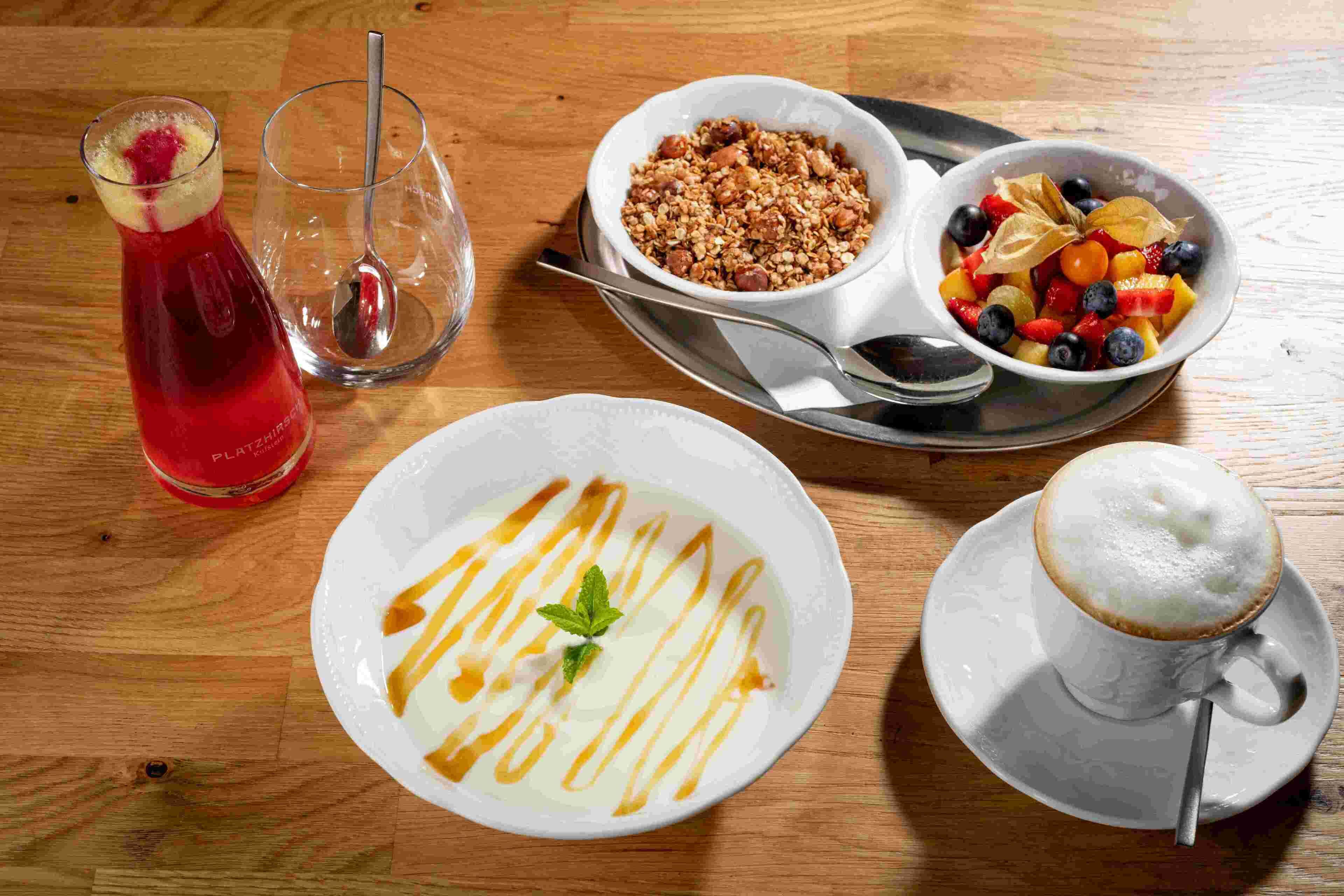 A breakfast setting with a red drink, a bowl of granola, a fruit bowl, a plate of yogurt with drizzles, and a cup of coffee.