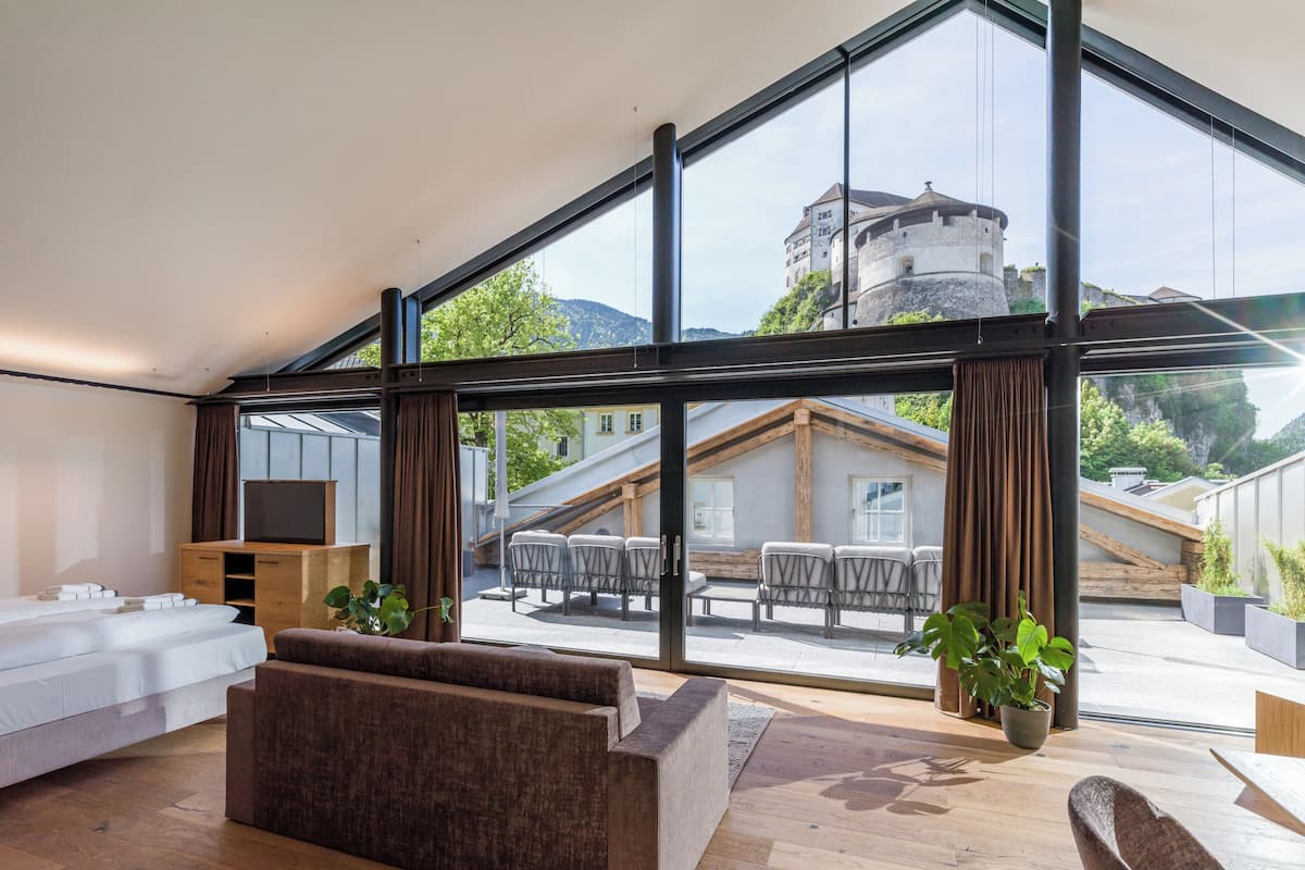 A modern hotel room with a large glass window, wooden flooring, a sofa, a wooden cabinet, and a view of a building with a tower.